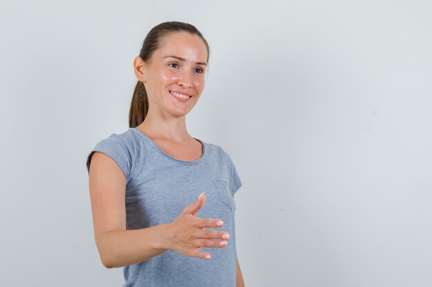 Junge Frau, die Hand für das Schütteln im grauen T-Shirt streckt und fröhlich, Vorderansicht schaut.