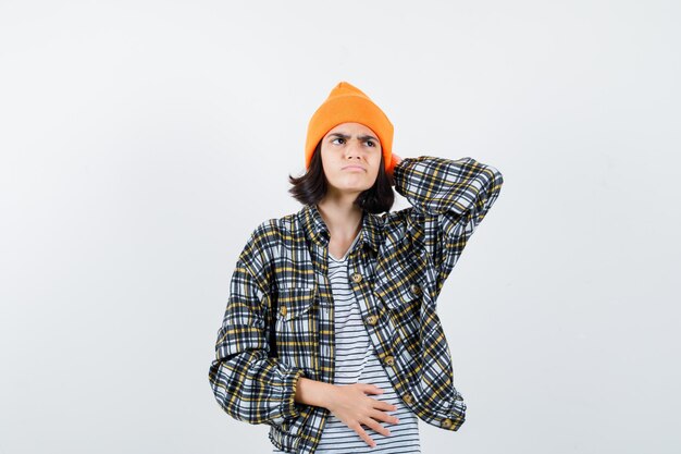 Junge Frau, die Hand hinter dem Kopf in einem orangefarbenen, karierten Hemd mit Hut hält, das verwirrt aussieht