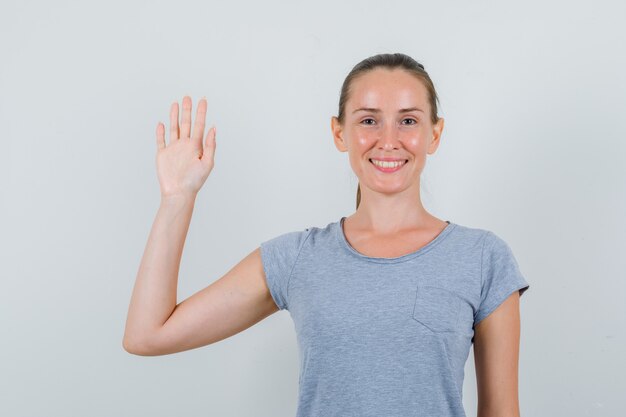 Junge Frau, die Hand im grauen T-Shirt winkt und fröhliche Vorderansicht schaut.