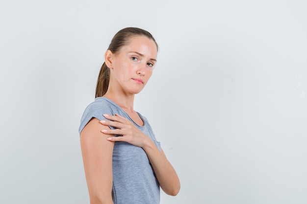 Junge Frau, die Hand nahe Brust in grauem T-Shirt hält und ernst schaut. Vorderansicht.