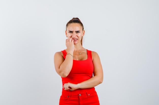 Junge Frau, die Hand nahe Mund in rotem Trägershirt, Hosen hält und Angst, Vorderansicht schaut.