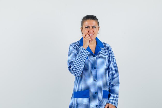 Junge Frau, die Hand nahe Mund setzt, über etwas im blauen Gingham-Pyjama-Hemd nachdenkt und überrascht schaut. Vorderansicht.