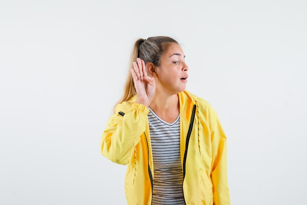 Junge Frau, die Hand nahe Ohr in T-Shirt, Jacke hält und neugierig schaut. Vorderansicht.