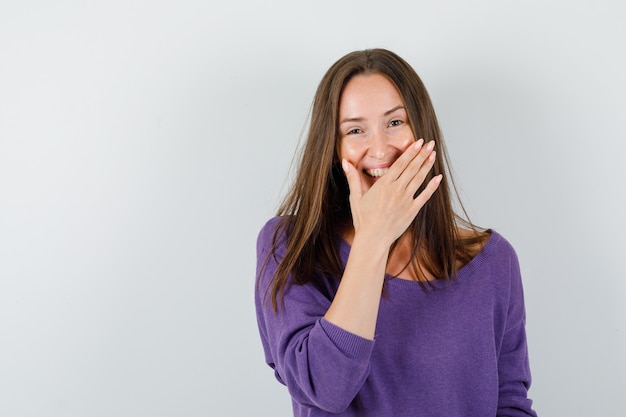 Junge Frau, die Hand über Mund im violetten Hemd hält und glückselig schaut. Vorderansicht.