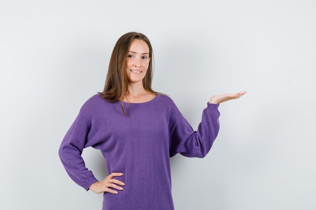Junge Frau, die Handfläche als etwas im violetten Hemd haltend und positiv aussehend hält. Vorderansicht.
