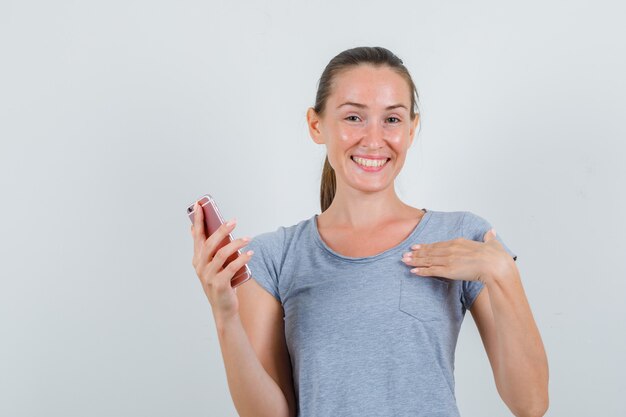 Junge Frau, die Handy hält, während sie sich im grauen T-Shirt zeigt und freudig schaut. Vorderansicht.