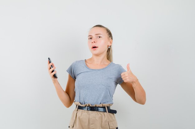 Junge Frau, die Handy hält, zeigt Daumen oben im T-Shirt, Vorderansicht der Hose.