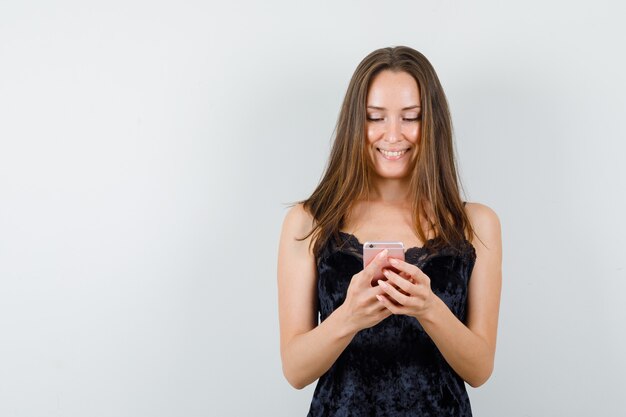 Junge Frau, die Handy im schwarzen Unterhemd benutzt und glücklich schaut