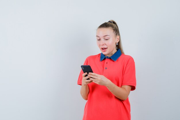 Junge Frau, die Handy im T-Shirt benutzt und glücklich schaut. Vorderansicht.