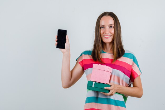 Junge Frau, die Handy und Geschenkboxen im T-Shirt hält und froh schaut, Vorderansicht.