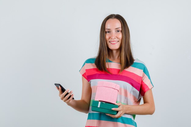 Junge Frau, die Handy und Geschenkboxen im T-Shirt hält und froh schaut, Vorderansicht.