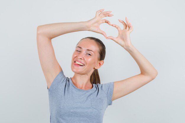 Junge Frau, die Herzgeste zeigt, zwinkert Auge in grauem T-Shirt, Vorderansicht.