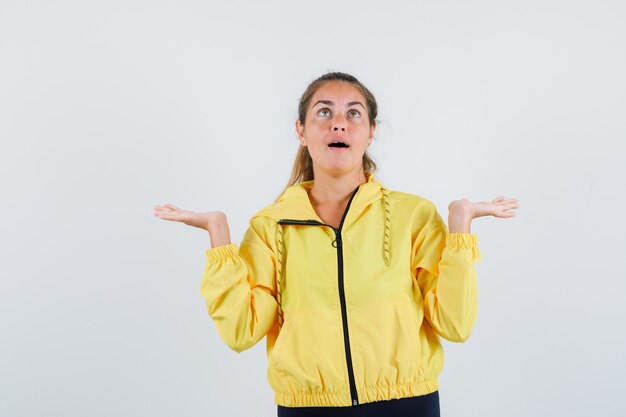 Junge Frau, die hilflose Geste zeigt, während sie im gelben Regenmantel aufschaut und verwirrt schaut