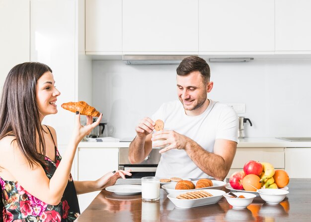 Junge Frau, die Hörnchen isst und ihr Ehemann, der Plätzchen in der Küche isst