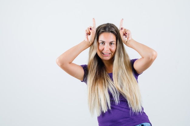 Junge Frau, die Hornzeichen auf ihrem Kopf im violetten T-Shirt zeigt und lustige Vorderansicht schaut.