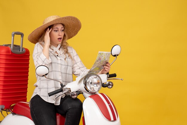 Junge Frau, die Hut trägt und auf Motorrad sitzt und Karte mit verwirrtem Gesichtsausdruck auf Gelb betrachtet