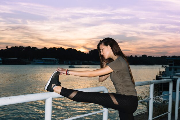 Junge Frau, die ihr Bein nahe Fluss bei Sonnenuntergang ausdehnt