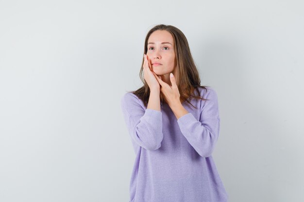 Junge Frau, die ihr Gesicht mit den Händen berührt, während sie in der lila Bluse aufwirft und vernünftig aussieht. Vorderansicht.