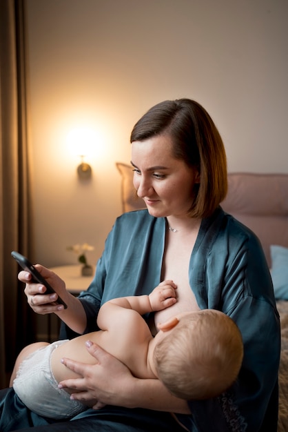 Junge Frau, die ihr süßes Baby stillt, während sie ihr Telefon überprüft