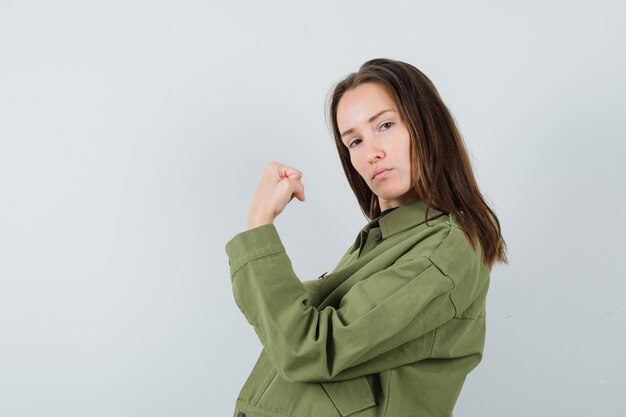 Junge Frau, die ihre Armmuskeln in der grünen Jacke zeigt und ernst schaut. Vorderansicht.