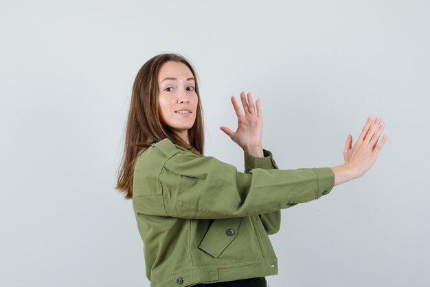 Junge Frau, die ihre Hände mit offenen Handflächen beiseite in grüner Jacke hebt und vorsichtig schaut. Vorderansicht.