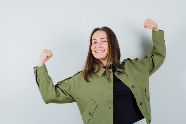 Junge Frau, die ihre Macht in der grünen Jacke zeigt und glückselige Vorderansicht schaut.