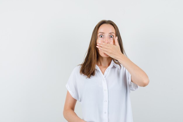 Junge Frau, die ihren Mund mit ihrer Hand in der weißen Bluse bedeckt und schockiert aussieht.