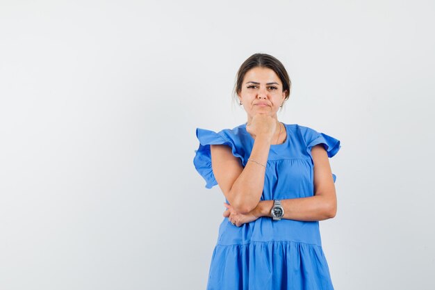 Junge Frau, die im blauen Kleid das Kinn auf die Faust stützt und vernünftig aussieht