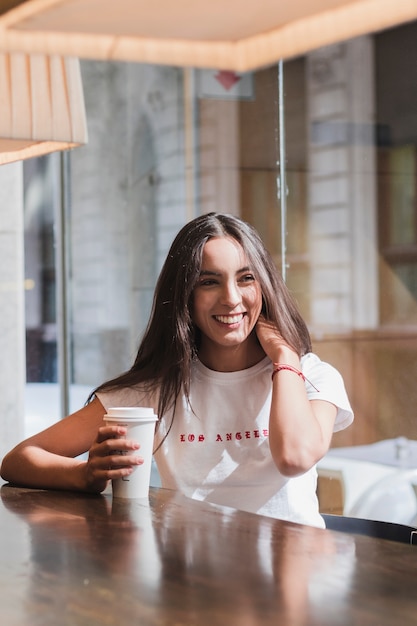 Junge Frau, die im Café bei Tisch hält Wegwerfkaffeetasse sitzt