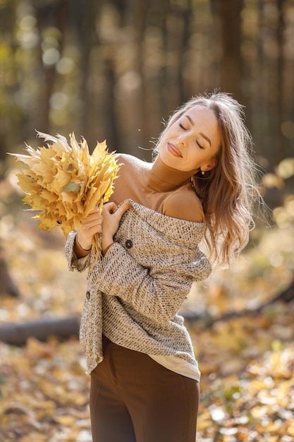 Junge Frau, die im Herbstwald steht. Brunettefrau, die gelbe Blätter hält. Mädchen, das Modebraunjacke trägt.