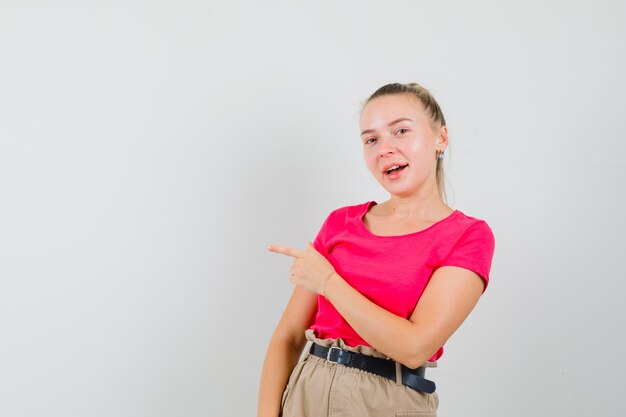 Junge Frau, die im T-Shirt, in der Hose und in der fröhlichen Vorderansicht beiseite zeigt.