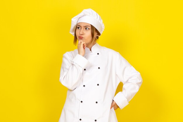 junge Frau, die im weißen Kochanzug und im weißen Kappen denkenden Ausdruck kocht
