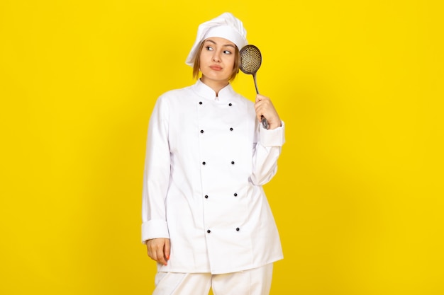 junge Frau, die im weißen Kochanzug und in der weißen Kappe kocht, die denkend hält, silbernen Löffel haltend