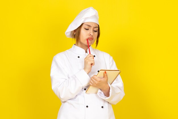 junge Frau, die im weißen Kochanzug und in der weißen Kappe kocht, die Notizen denkend aufschreiben