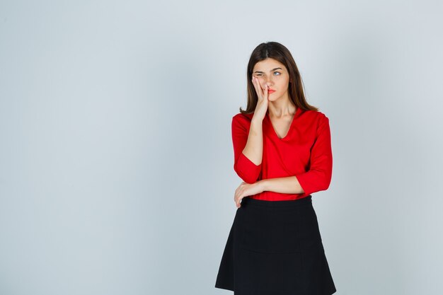 Junge Frau, die in denkender Haltung steht und Hand nahe Mund in rote Bluse setzt