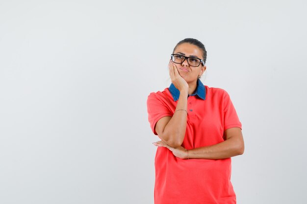 Junge Frau, die in denkender Haltung steht, Wange auf Handfläche im roten T-Shirt und in der Brille lehnt und nachdenklich schaut. Vorderansicht.