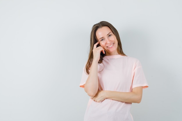 Junge Frau, die in der denkenden Haltung im rosa T-Shirt steht und fröhlich schaut. Vorderansicht.