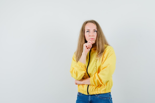 Junge Frau, die in der denkenden Haltung in der gelben Bomberjacke und in der blauen Jeans steht und nachdenklich, Vorderansicht schaut.