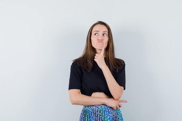 Junge Frau, die in der denkenden Haltung steht und in schwarzem T-Shirt und blauem Rock wegschaut und nachdenklich schaut