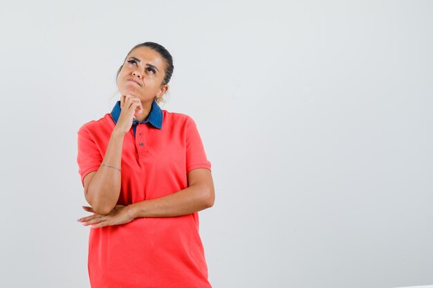 Junge Frau, die in der denkenden Pose im roten T-Shirt steht und nachdenklich, Vorderansicht schaut.