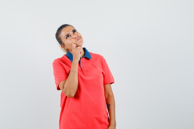 Junge Frau, die in der denkenden Pose im roten T-Shirt steht und nachdenklich, Vorderansicht schaut.