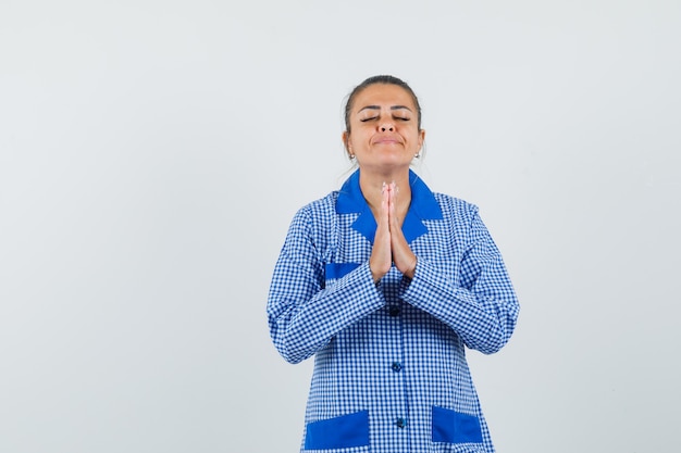 Junge Frau, die in der Gebetshaltung im blauen Gingham-Pyjamahemd steht und entspannt, Vorderansicht schaut.