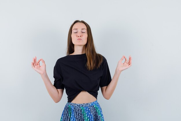 Junge Frau, die in der Meditationsgeste steht und Küsse an Kamera in schwarzem T-Shirt und blauem Rock sendet und attraktiv aussieht