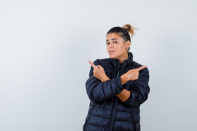 Junge frau, die in pufferjacke nach links und rechts zeigt und unentschlossen aussieht. vorderansicht.