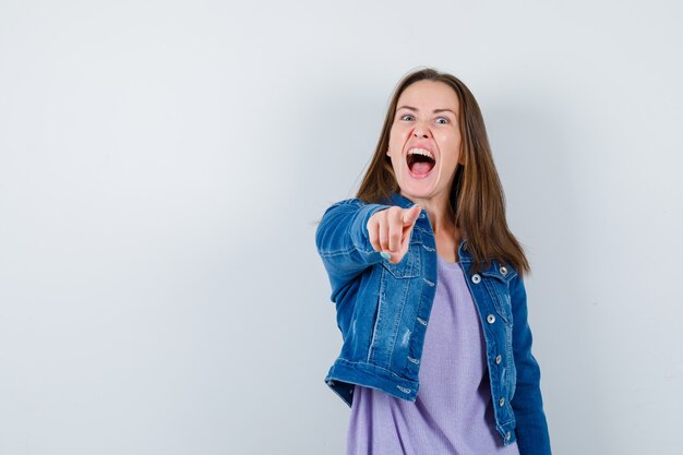 Junge Frau, die in T-Shirt, Jacke nach vorne zeigt und verrückt aussieht. Vorderansicht.