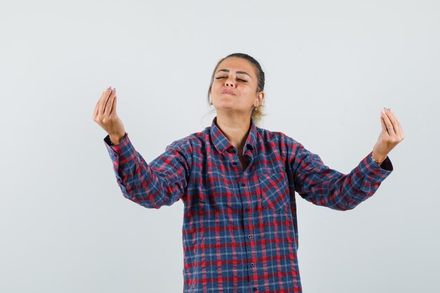 Junge Frau, die italienische Geste im karierten Hemd zeigt und ruhige Vorderansicht schaut.