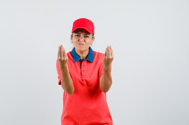 Junge Frau, die italienische Geste im roten T-Shirt und in der Mütze zeigt und erfreut schaut. Vorderansicht.