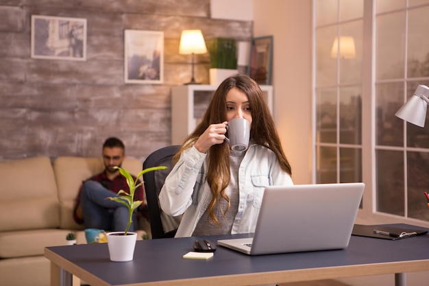 Junge Frau, die Kaffee trinkt und am Laptop von zu Hause aus arbeitet. Freund sitzt bequem auf dem Sofa im Hintergrund