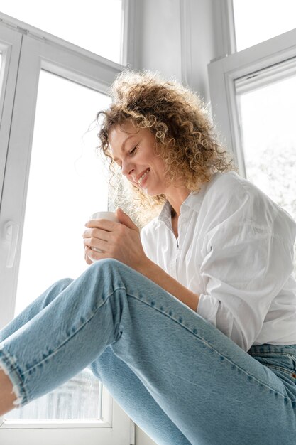 Junge Frau, die Kaffee zu Hause nahe dem Fenster trinkt