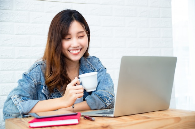 Junge Frau, die Kaffeetasse und Gebrauchslaptop im Büro hält